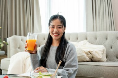 Çekici, gülümseyen genç bir Asyalı kadın oturma odasındaki kahve masasında otururken elinde bir bardak portakal suyu tutarak kameraya gülümsüyor. İnsanlar ve sağlıklı gıda kavramları