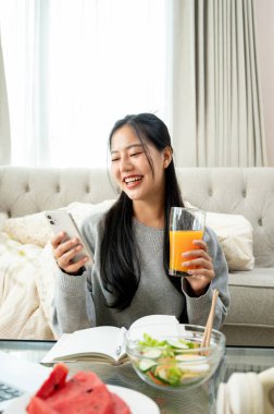 Güzel, neşeli genç bir Asyalı kadın telefonuyla sohbet ederken gülüyor ve oturma odasındaki kahve masasında sağlıklı bir kahvaltı yapıyor. insanlar ve ev hayatı kavramları