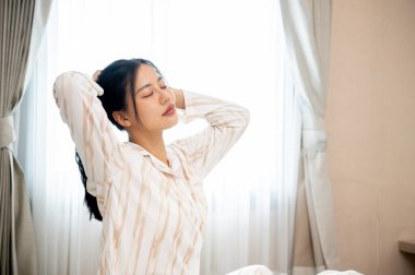 Güzel, rahat, pijamalı bir Asyalı kadın sabah yatak odasında uyandıktan sonra yatağında saçlarını topluyor. insanlar ve ev hayatı kavramları