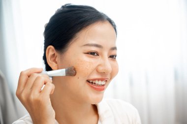 Güzel, gülümseyen Asyalı bir kadının yakın plan görüntüsü yüzüne bir fırça ile bir yüz tedavi maskesi uyguluyor. Cilt bakımı, güzellik, kozmetik, kişisel bakım.