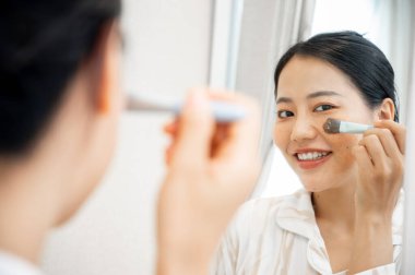 Pijamalı genç ve güzel bir Asyalı kadın yüz tedavi maskesini aynanın önünde fırçasıyla uyguluyor..