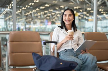 Kendine güvenen Asyalı bir kadın, elinde kahve fincanı ve dijital tabletle havaalanı terminalinde bir bekleme koltuğunda oturur. Yatma zamanını bekler, gülen yüzüyle başka yere bakar..