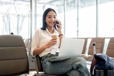 Kendine güvenen Asyalı bir bayan yolcu telefonla konuşuyor ve havaalanı terminalindeki bekleme koltuğunda dizüstü bilgisayarıyla uçağını bekliyor. Uzaktan çalışma, ulaşım.