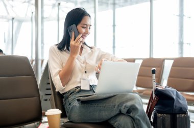 Kendine güvenen Asyalı bir iş kadını telefonda konuşurken ve havaalanı terminalinde dizüstü bilgisayarıyla çalışırken kol saatini kontrol ediyor ve uçuşunu bekliyor. Uzaktan çalışıyor.