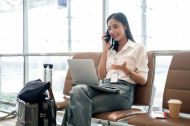 Kendine güvenen Asyalı bir bayan yolcu telefonla konuşuyor ve havaalanı terminalindeki bekleme koltuğunda dizüstü bilgisayarıyla uçağını bekliyor. Uzaktan çalışma, ulaşım.