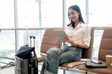 Çekici bir Asyalı bayan yolcu, havaalanı terminalinde uçağını beklerken dijital tabletini kullanarak uzaktan çalışıyor. İnsanlar, yaşam tarzı ve ulaşım