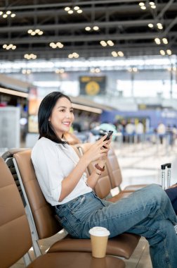 Çekici bir Asyalı bayan yolcu havaalanı terminalinde bekleme koltuğunda oturuyor ve akıllı telefonunu kullanarak uçuşunu bekliyor. İnsanlar, seyahat, ulaşım, yaşam tarzı