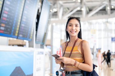 Çekici, gülümseyen Asyalı genç bir bayan turist havaalanı terminalinde duruyor, uçuş bilgi ekranının önünde, yazın seyahat ediyor. insanlar ve ulaşım kavramları