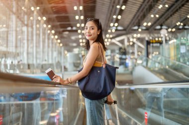 Güzel bir Asyalı genç bayan turist bavuluyla havaalanı yürüyen merdiveninde kameraya gülümsüyor, uçakla seyahat ediyor. insanlar ve ulaşım kavramları, kopyalama alanı ile