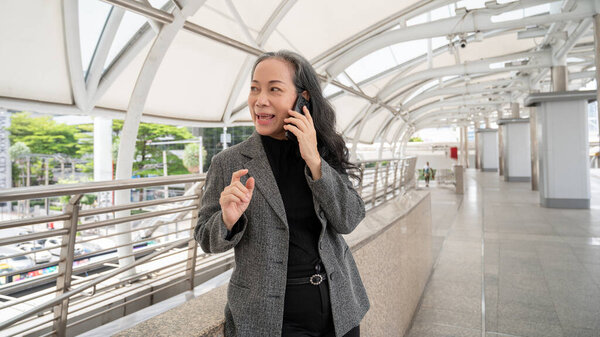 Уверенная в себе зрелая азиатская бизнесвумен в костюме разговаривает по телефону со своим клиентом, стоя на тротуаре в городе. Предприниматели и концепции беспроводных технологий