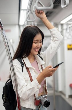 Güzel, gülümseyen, kameralı genç bir Asyalı turist elinde bir tırabzan tutuyor ve akıllı telefonunu bir yerlere giderken kullanıyor, bir şehir merkezinde toplu taşıma aracını kullanıyor..