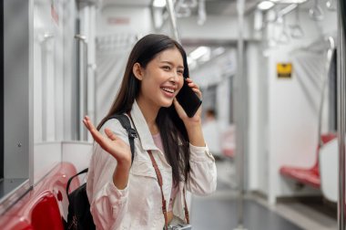 Sırt çantalı, mutlu, genç Asyalı bir turist şehir içi seyahatine çıkan bir trenle ya da metroyla telefonda konuşuyor. insanlar ve ulaşım kavramları
