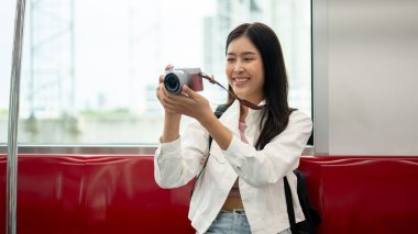 Neşeli, çekici, genç Asyalı bir bayan turist kamerasıyla fotoğraf çekerken bir gökyüzü treniyle gidip gelirken, toplu taşımacılığın keyfini çıkarıyor..