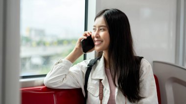 Sırt çantalı mutlu, çekici bir Asyalı kadın dışarıdaki manzaraya bakıyor ve gökyüzü trenine binerken telefonla konuşuyor. İnsanlar, şehir hayatı, yaşam tarzı, ulaşım