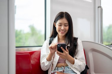 Sırt çantası ve kamerası olan güzel, gülümseyen Asyalı genç bir bayan gezgin gökyüzü trenine binerken akıllı telefonunu kullanıyor, sohbet ediyor ya da mesajlar okuyor. insanlar ve ulaşım