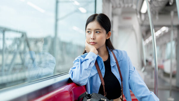 A positive, thoughtful young Asian female tourist passenger is thinking about something and gazing at the view outside while commuting in the city by sky train. traveling, public transport