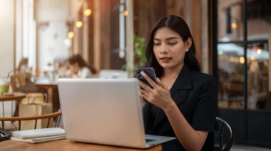 Siyah takım elbiseli çekici bir Asyalı iş kadını akıllı telefonunu kullanıyor, mesajları okuyor, dizüstü bilgisayarı üzerinde çalışıyor ve şehirdeki bir kafede uzaktan çalışıyor..