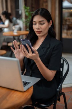 Siyah takım elbiseli çekici bir Asyalı iş kadını akıllı telefonunu kullanıyor, mesajları okuyor, dizüstü bilgisayarı üzerinde çalışıyor ve şehirdeki bir kafede uzaktan çalışıyor..