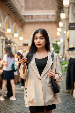 Çekici, kendine güveni tam bir Asyalı kadın şehirdeki bir alışveriş caddesinde duruyor, akıllı telefonunu tutuyor ve kameraya bakıyor..