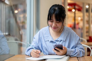 Genç bir Asyalı kadın ödevlerini yaparken akıllı telefonunu kullanıyor ya da bir kitaba not alıyor, bir kahve dükkanında masada oturuyor. İnsanlar ve kablosuz teknoloji kavramları