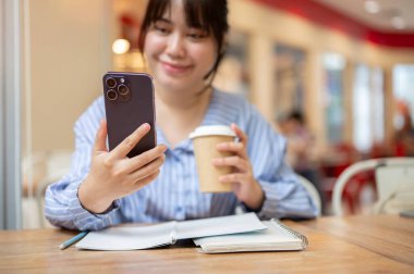 Seçici bir odak noktası, mutlu bir Asyalı kadın telefonuna bakıyor, mesajlar okuyor veya kafede otururken videolar izliyor. İnsanlar ve kablosuz teknoloji kavramları