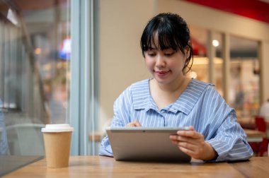 Çekici genç bir Asyalı kadın dijital tabletini bir kafede kullanıyor, tablet üzerinde resim yazıyor ya da dizayn ediyor, uzaktan çalışıyor. kablosuz teknoloji