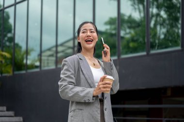 Kendine güvenen, takım elbiseli güzel Asyalı bir iş kadını elinde kahve fincanıyla şehirde yürürken telefonda konuşuyor. modern şehir hayatı ve iş insanları kavramları