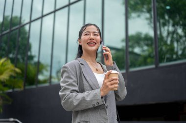 Kendine güvenen, takım elbiseli güzel Asyalı bir iş kadını elinde kahve fincanıyla şehirde yürürken telefonda konuşuyor. modern şehir hayatı ve iş insanları kavramları