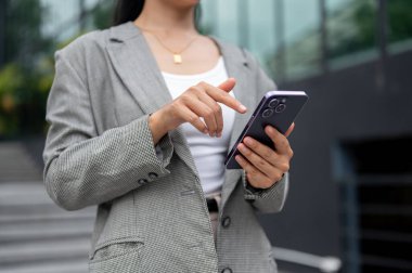 Gri takım elbiseli Asyalı bir iş kadını şehirde yürürken mesaj okuyor ya da akıllı telefonundan mesajlara cevap veriyor. kablosuz teknoloji ve şehir hayatı kavramları