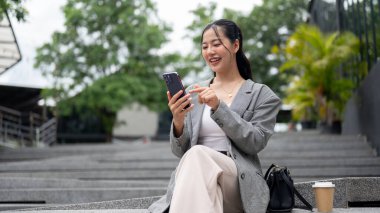 Mutlu, güzel bir Asyalı iş kadını şehrin dışındaki merdivenlerde otururken mesaj okuyor ya da akıllı telefonuyla sohbet ediyor. kablosuz teknoloji ve insanların kavramları