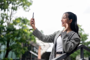 Gri takım elbiseli, olumlu, güzel bir Asyalı iş kadını şehirde dışarıda dikilirken akıllı telefonuyla el sallıyor ve bir video görüşmesinde konuşuyor. İnsanlar ve kablosuz teknoloji