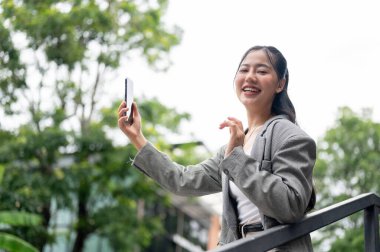 Gri takım elbiseli, Asyalı olumlu, güzel bir iş kadını dışarıda akıllı telefonuyla konuşurken kameraya bakıp gülümsüyor. İnsanlar ve kablosuz teknoloji
