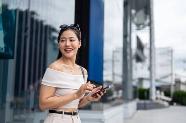 Şık giyinmiş güzel, gülümseyen Asyalı bir kadın elinde akıllı bir telefonla alışveriş merkezinin önünde kameradan uzağa bakıyor..