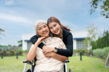 Sevimli bir Asyalı torun, tekerlekli sandalyede olan büyükannesine sarılıyor. Onu parkta gezdirirken, birlikte vakit geçirirken, sevgi ve özenle kucaklaşırken ve ikisi de kameraya gülümserken..