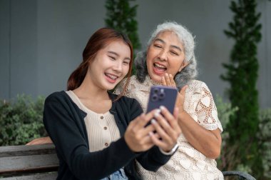 Mutlu bir emekli Asyalı büyükanne telefonda sosyal medyada videoları izlerken ve bahçede bir bankta dinlenirken torunuyla gülüyor ve eğleniyor..
