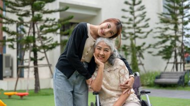 Sevimli bir Asyalı torun, tekerlekli sandalyede olan büyükannesine sarılıyor. Onu parkta gezdirirken, birlikte vakit geçirirken, sevgi ve özenle kucaklaşırken ve ikisi de kameraya gülümserken..