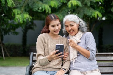 Şefkatli, mutlu bir Asyalı torun büyükannesine akıllı telefonuyla bir video gösteriyor, parkta bir bankta dinlenirken konuşmaktan keyif alıyor. insanlar ve teknoloji kavramları