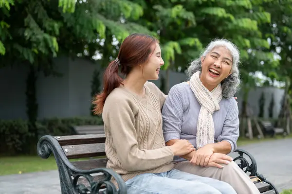 Güzel bir Asyalı torun ve mutlu bir emekli büyükanne parkta bir bankta dinlenirken gülüyor ve sohbet ediyorlar. Mutlu an, aile bağları, boş zaman.