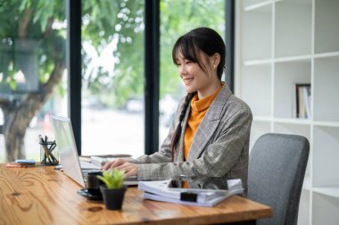 Çekici, pozitif Asyalı bir ofis çalışanı ofisteki masasında dizüstü bilgisayarı üzerinde çalışıyor, klavyesinde yazıyor, internette geziniyor. İş insanları kavramı