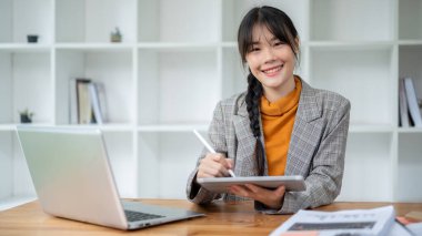 Moda kıyafeti giymiş çekici bir Asyalı iş kadını elinde dijital tabletle masasında oturmuş kameraya gülümsüyor. İş insanları ve kablosuz teknoloji kavramları
