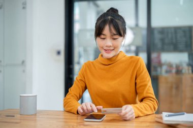 Güzel, mutlu, genç Asyalı bir kadın kredi kartı tutuyor ve akıllı telefonunu kullanıyor, internet faturalarını ödemek için mobil bankacılık uygulamasını kullanıyor. Nakitsiz, çevrimiçi alışveriş, online ödeme, para transferi