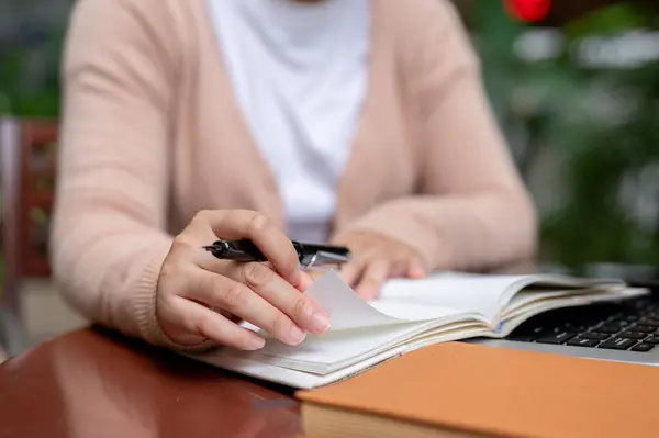 Stock image A close-up image of a woman in casual clothes holding a pen, writing something in a book, studying on lecturing the online class, doing homework, sitting at a table outdoors.