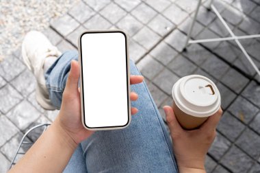 Kot pantolonlu bir kadının elinde kahve fincanıyla dışarıda otururken ve akıllı telefonunu kullanırken kafeteryada takılırken en güzel görüntüsü. Beyaz ekran modeli olan bir akıllı telefon..