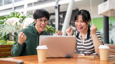 Two cheerful and excited young Asian friends, male and female, are looking at a laptop screen, surprised by the good news and celebrating together while sitting at an outdoor table.