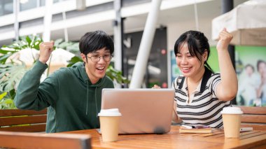 İki neşeli ve heyecanlı Asyalı genç arkadaş, erkek ve kadın, bir dizüstü bilgisayara bakıyorlar, iyi habere şaşırıyorlar ve birlikte kutluyorlar açık bir masada otururken..