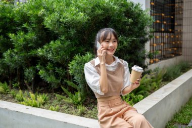 Çekici, mutlu bir Asyalı kadın öğle yemeği molasında dışarıda dinlenirken kahve içiyor ve konuşuyor..