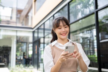 Sıradan giysiler içindeki çekici, mutlu, genç Asyalı bir kadın bina alanında dikilirken akıllı telefonunu kullanıyor. sohbet, telefonda kaydırma, uygulamalar, kablosuz teknoloji ve yaşam tarzı