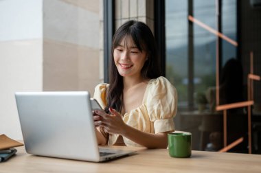 Güzel, gülümseyen Asyalı bir kadın masasında akıllı telefonunu kullanıyor, çalışırken, ofisinde otururken....