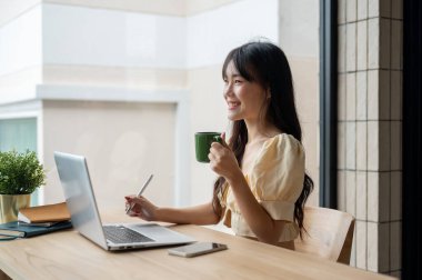 Genç ve güzel bir Asyalı kadın ofisteki masasında laptopuyla çalışırken kahve içiyor..