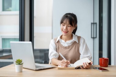 Güzel bir Asyalı kız üniversite öğrencisi dizüstü bilgisayarıyla bir ders çalışırken bir kitapta ders veriyor, bir odada oturuyor..